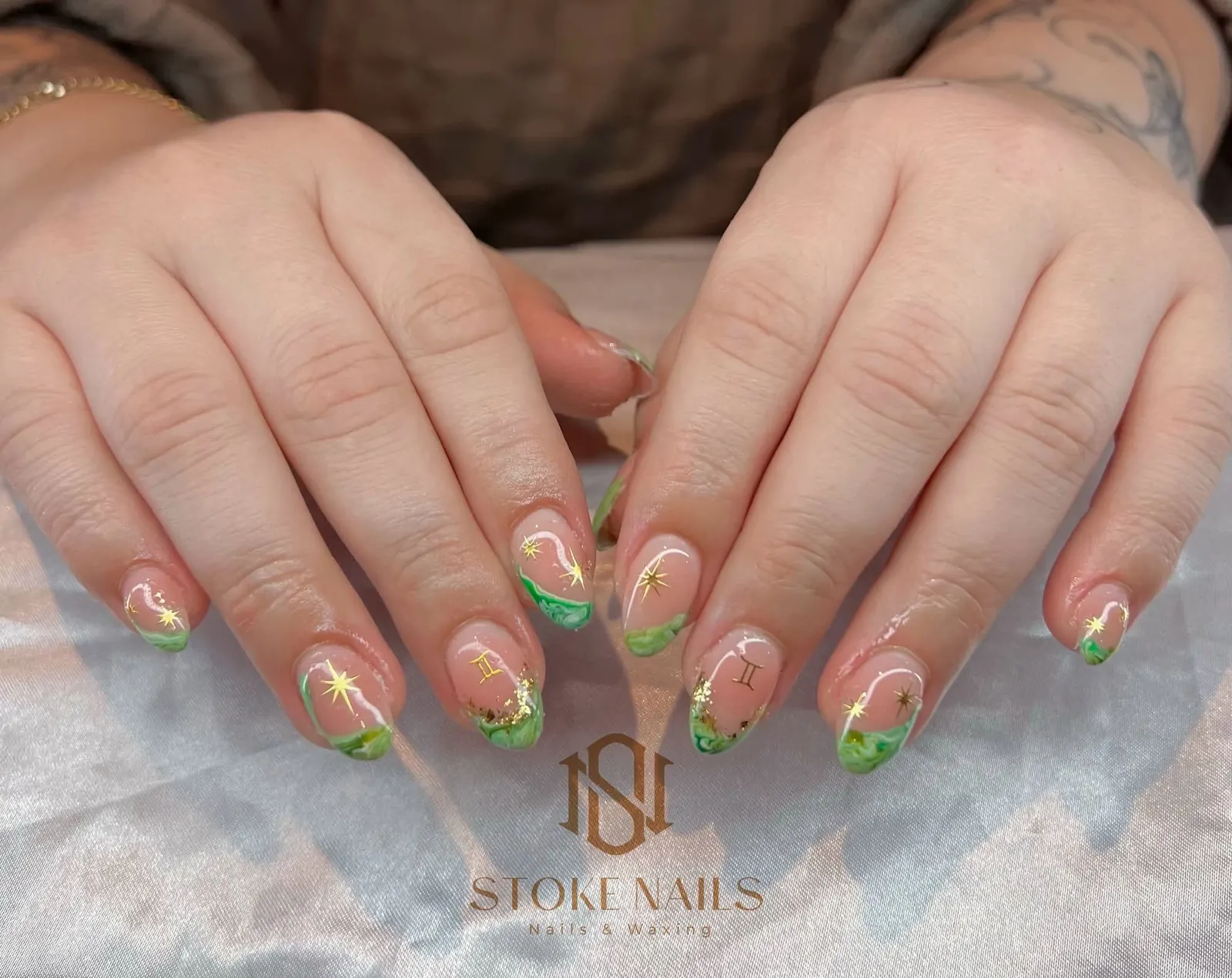 Both hands resting on a pale cloth, showing sheer nude nails with green tips and small gold star details