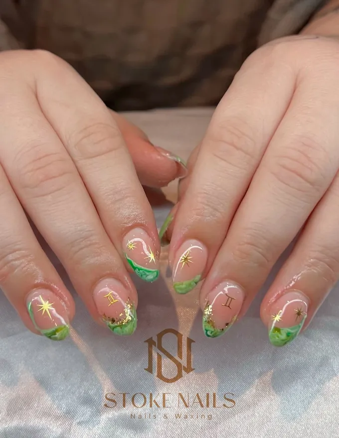 Two hands with almond nails in a sheer nude base, finished with green tips and fine gold detailing