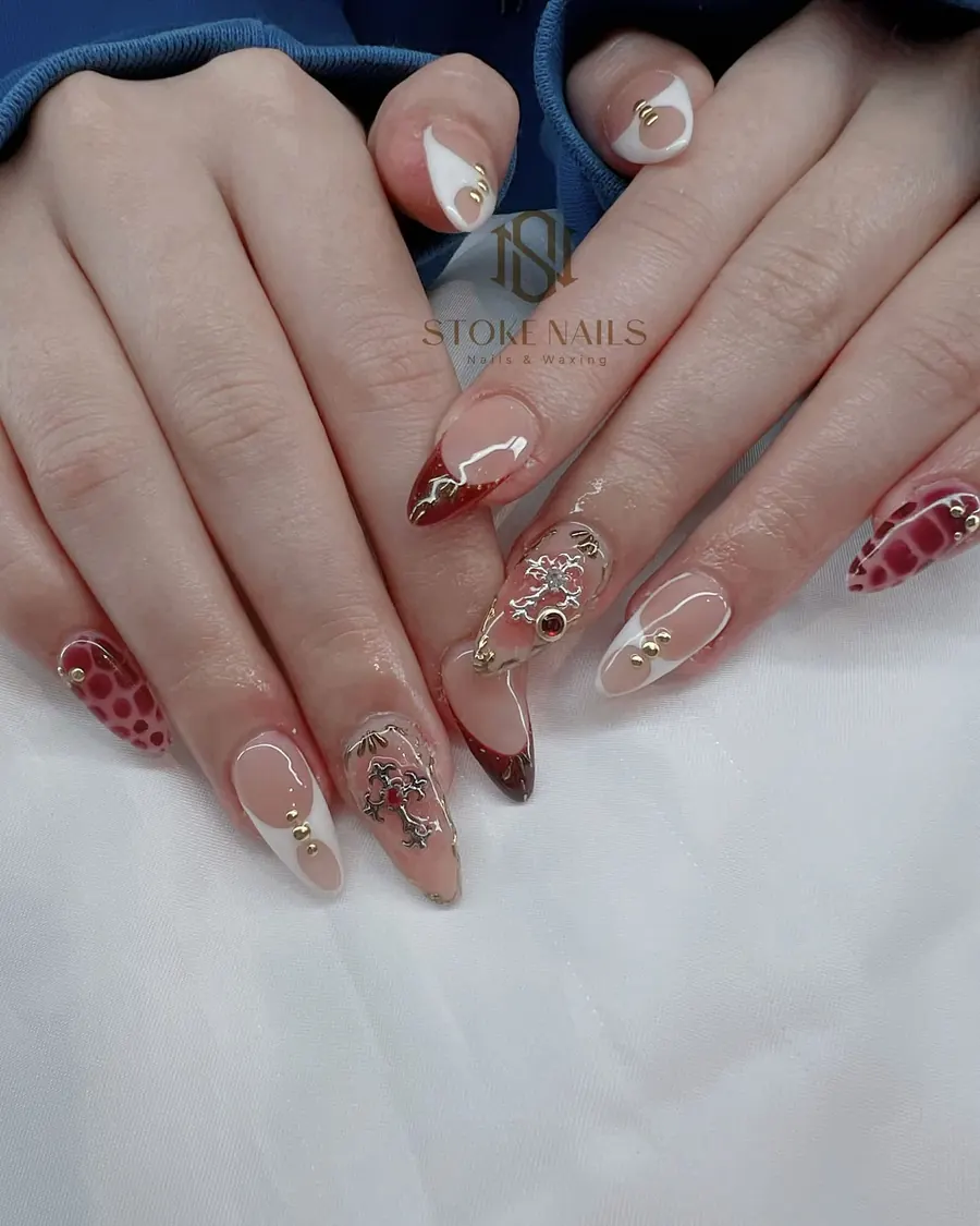 Long almond nails mixing red French tips, red patterned accents and gold charms on a nude base